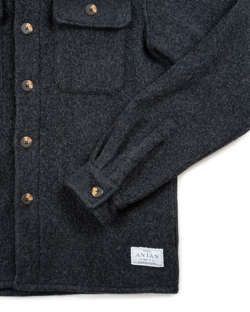 Close-up of a dark charcoal wool overshirt jacket with bronze buttons and a white Antan brand label near the hem, shown on a white background.