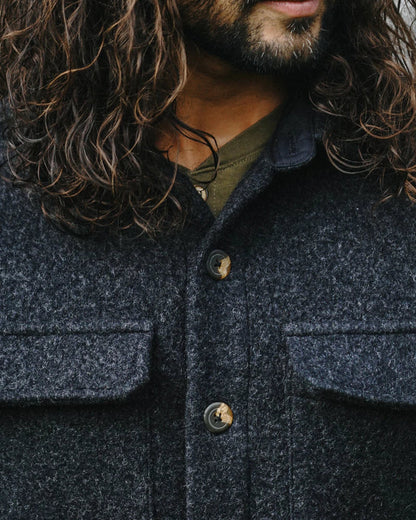 Close-up of a person wearing a dark charcoal gray textured wool-blend button-up jacket with brown buttons, layered over an olive green shirt. The image features long, wavy brown hair and a beard.