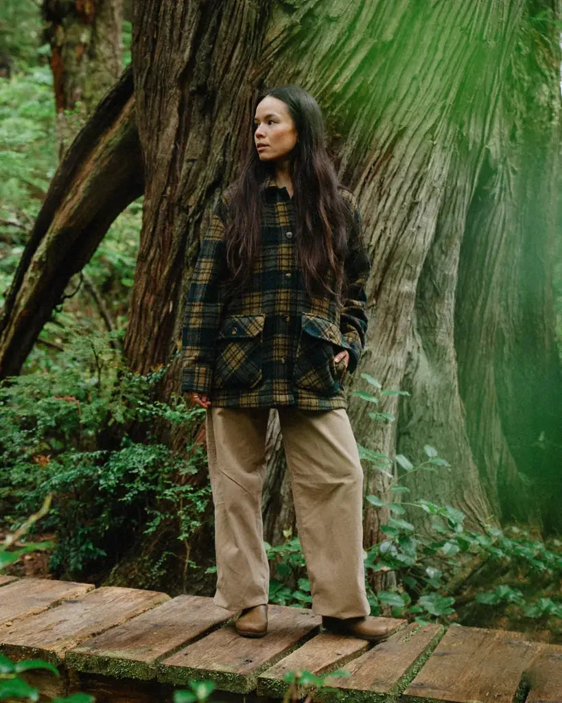 Person standing on a wooden platform in a forest with a large tree behind them wearing a plaid wool coat