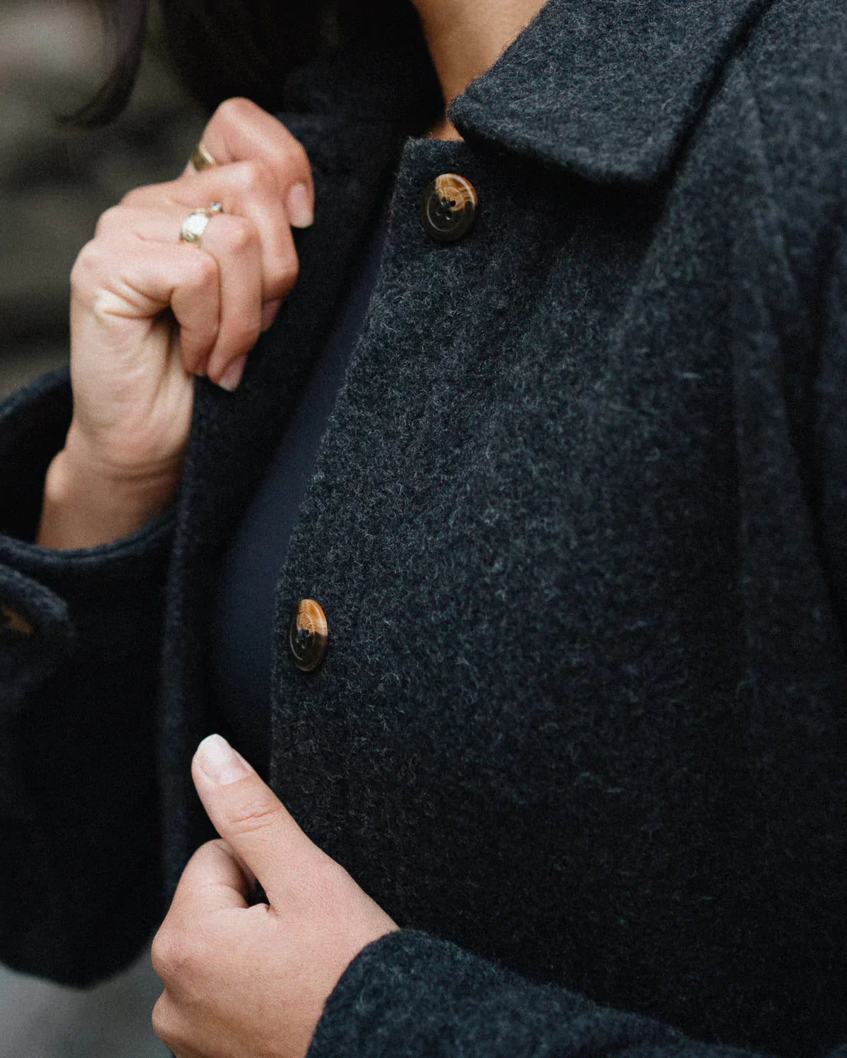 Close-up of a person wearing a dark charcoal gray wool-blend coat with brown buttons, holding the collar with one hand and adjusting the front with the other.