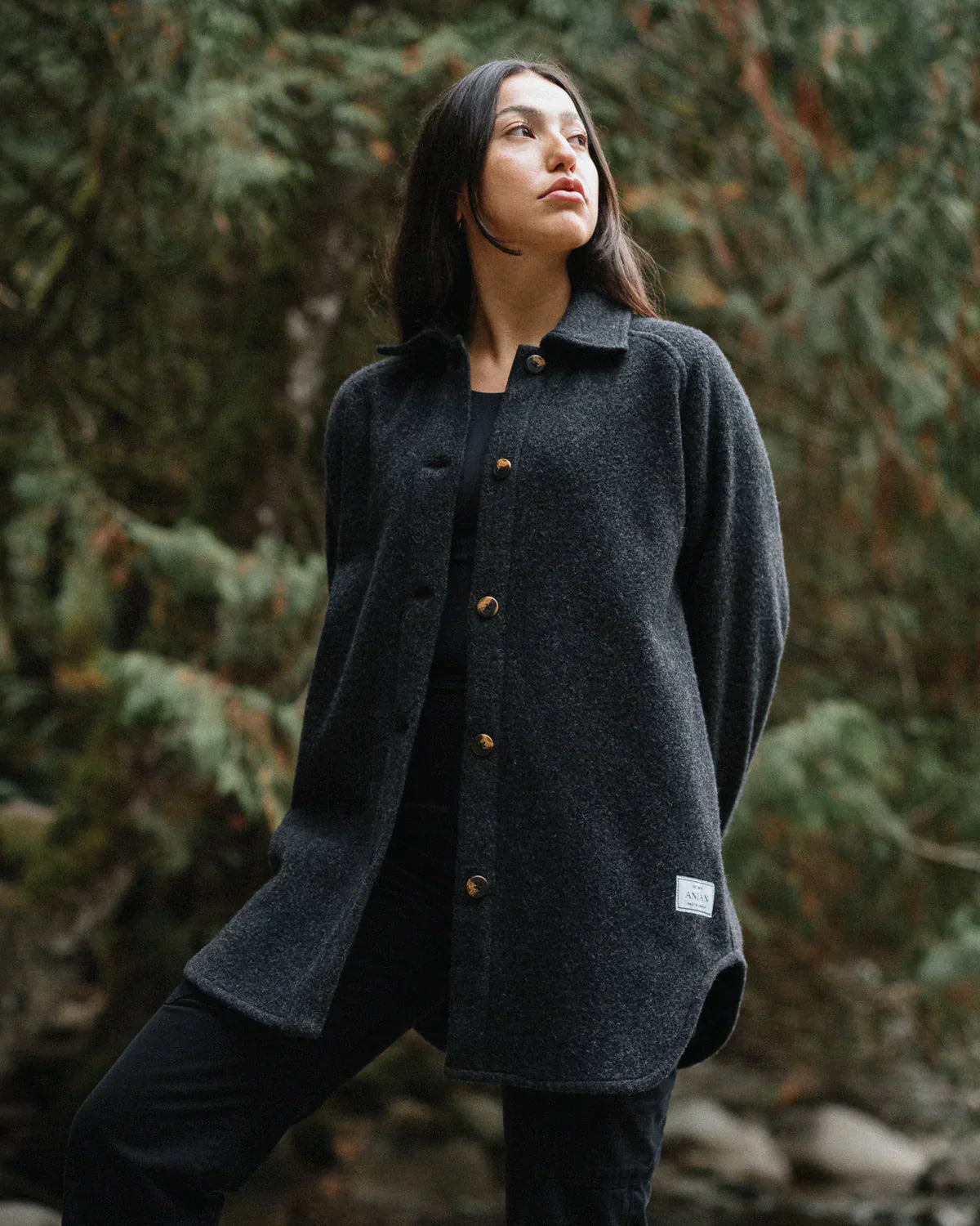 A woman wearing a dark charcoal gray textured button-up coat with brown buttons, standing outdoors in a forest setting.