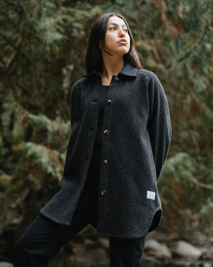 A woman wearing a dark charcoal gray textured button-up coat with brown buttons, standing outdoors in a forest setting.