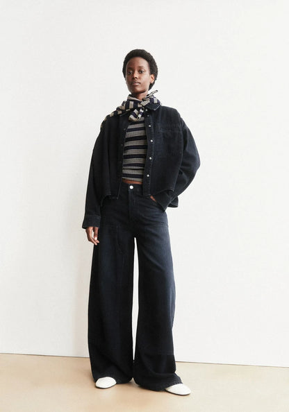 Person wearing a dark jacket, striped shirt, and wide-leg pants against a white background