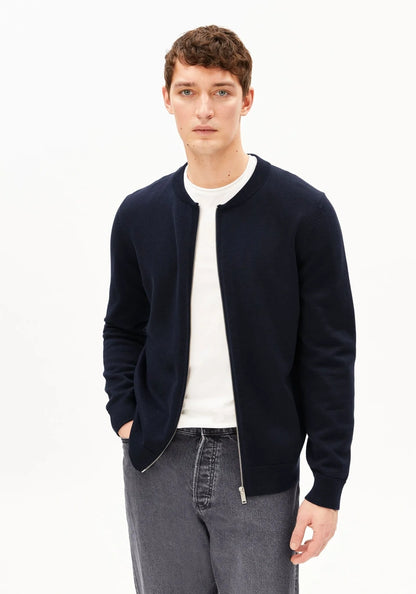 A man wearing a navy blue zip-up knit cardigan over a white t-shirt and gray jeans, standing against a plain white studio background.
