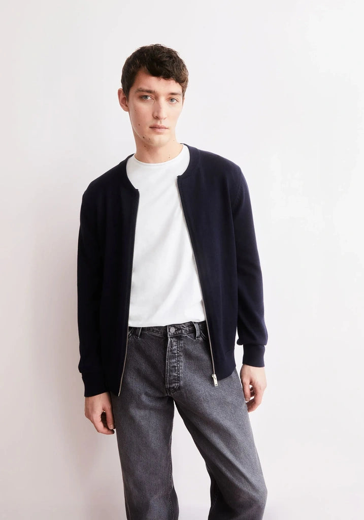 A young man modeling a dark navy blue zip-up knit cardigan over a white crewneck t-shirt and gray jeans, standing against a plain off-white studio background.