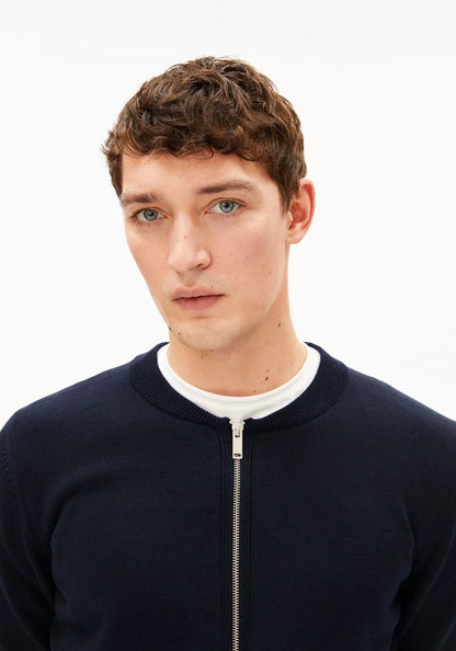 A close-up portrait of a young man with short brown hair wearing a dark navy ribbed zip-up sweater over a white crew-neck t-shirt, set against a plain white studio background.