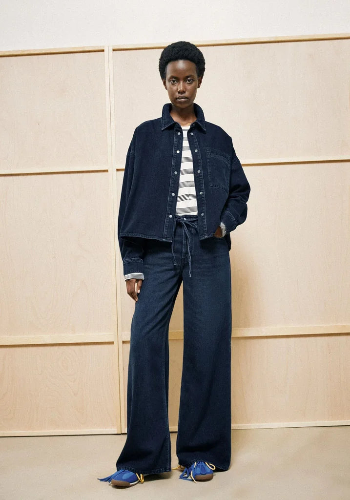 A model stands against a light wood-paneled wall wearing a dark indigo denim outfit consisting of a cropped button-up shirt, matching wide-leg pants, and a striped t-shirt underneath, paired with blue slip-on shoes.