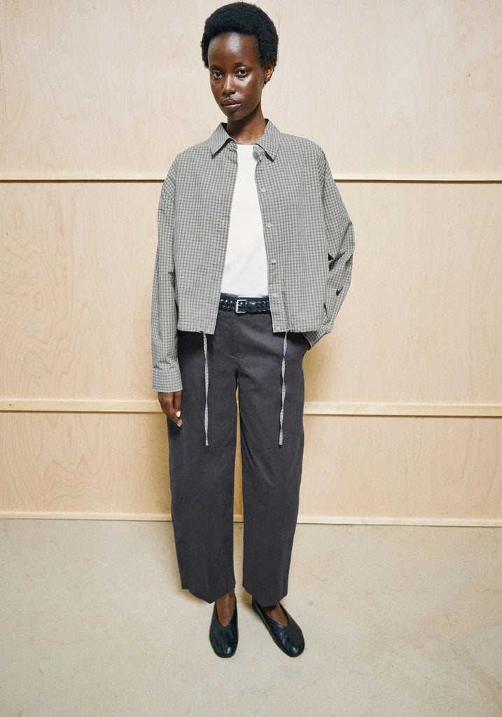 A model stands against a light wood-paneled wall wearing a cropped black and white gingham jacket over a white t-shirt, dark gray wide-leg trousers, and black leather loafers.
