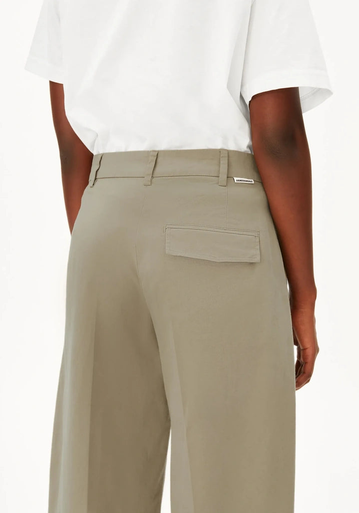 A close-up back view of a model wearing high-waisted, wide-leg trousers in a light khaki color, paired with a white t-shirt. The trousers feature a belt loop, a welt pocket on the right side, and a small silver brand tag on the back waistband.