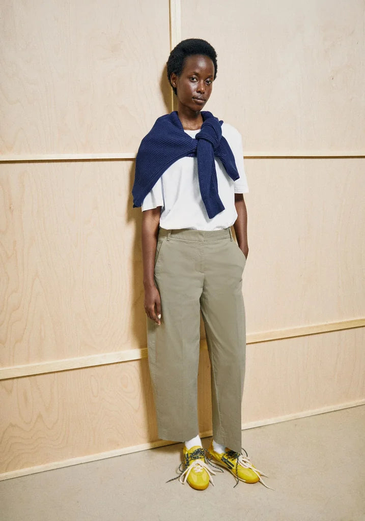 A model stands against a light wood-paneled wall wearing a white short-sleeve top with a navy blue draped scarf, beige wide-leg cropped trousers, and bright yellow sneakers.
