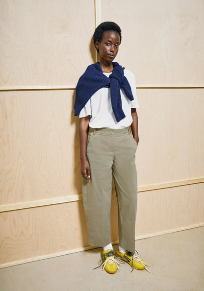 A model stands against a light wood-paneled wall wearing a white short-sleeve top with a navy blue draped scarf, beige wide-leg cropped trousers, and bright yellow sneakers.
