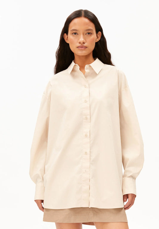 Woman wearing the ARMEDANGELS Canada Barrel Sleeve Poplin Blouse in pale peach buttoned and untucked over a beige mini skirt, front view against a white background