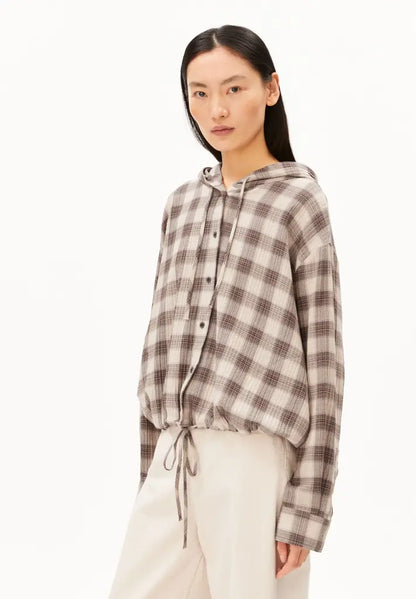 A model wearing a brown and beige plaid hooded button-up shirt with a drawstring hem, paired with cream-colored pants, against a plain white studio background.
