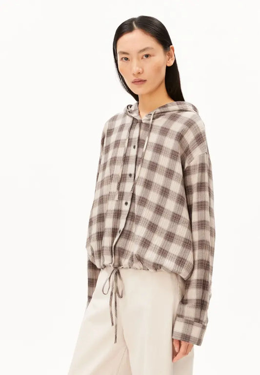 A model wearing a brown and beige plaid hooded button-up shirt with a drawstring hem, paired with cream-colored pants, against a plain white studio background.