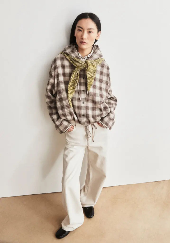A model stands against a plain off-white wall wearing a brown and beige plaid hooded shirt with a gold patterned scarf, cream wide-leg trousers, and black shoes.