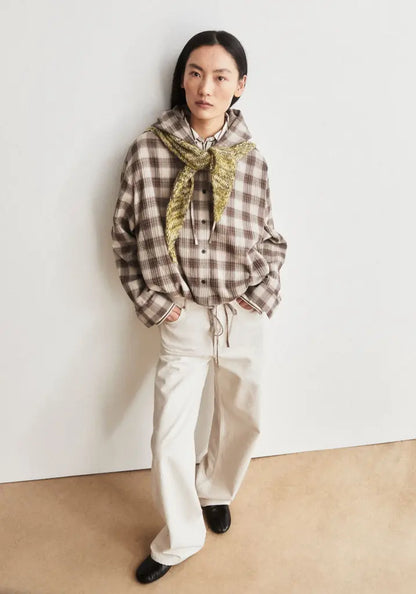 A model stands against a plain off-white wall wearing a brown and beige plaid hooded shirt with a gold patterned scarf, cream wide-leg trousers, and black shoes.
