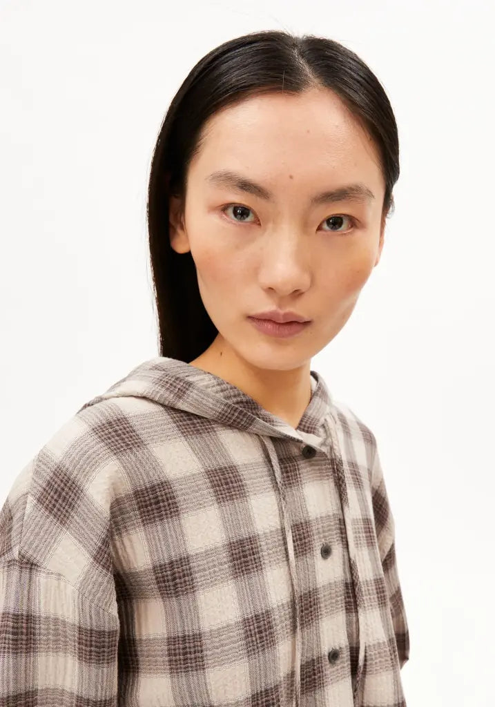 A model with long dark hair wearing a brown and beige plaid hooded button-up shirt, photographed against a plain white background.