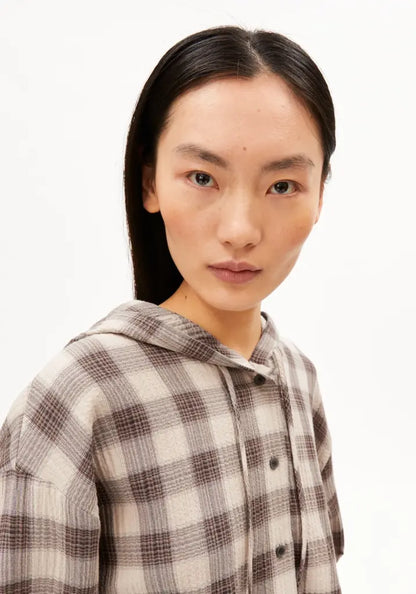A model with long dark hair wearing a brown and beige plaid hooded button-up shirt, photographed against a plain white background.
