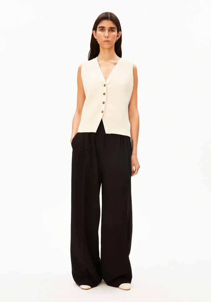 Full-length front view of a woman wearing the ARMEDANGELS Canada Compact Buttoned Knit Vest in oatmilk with black wide-leg trousers against a white background.