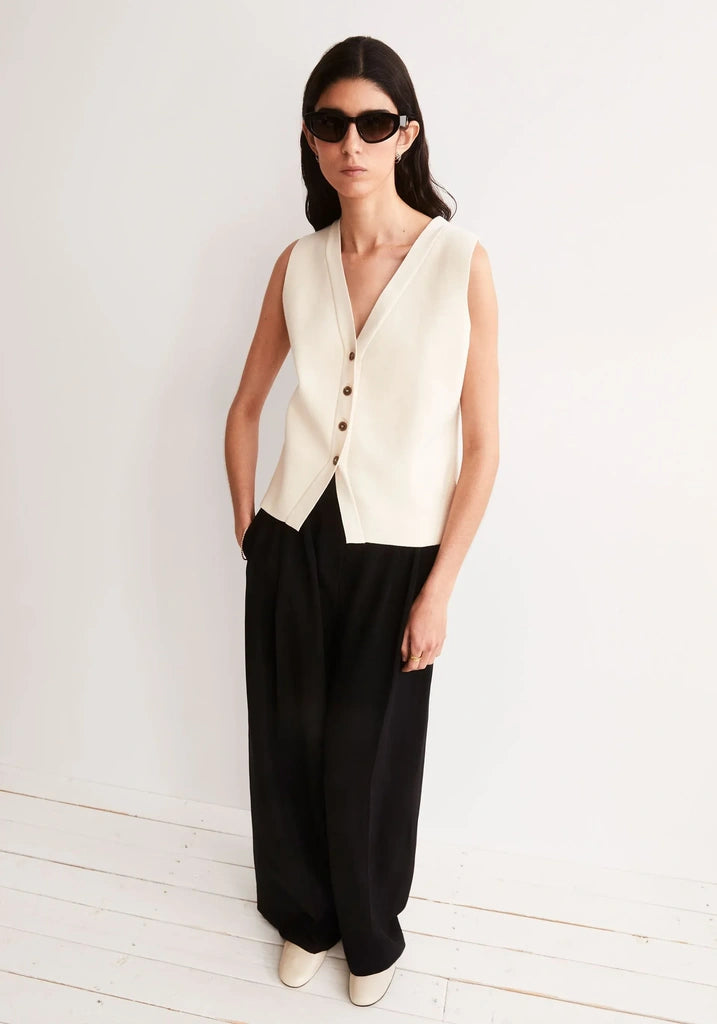 Woman in sunglasses wearing the ARMEDANGELS Canada Compact Buttoned Knit Vest in oatmilk with black wide-leg trousers, standing on a white wooden floor against a pale wall.