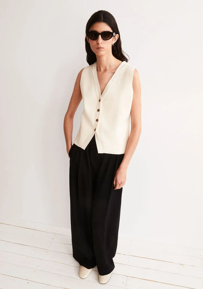 Woman in sunglasses wearing the ARMEDANGELS Canada Compact Buttoned Knit Vest in oatmilk with black wide-leg trousers, standing on a white wooden floor against a pale wall.