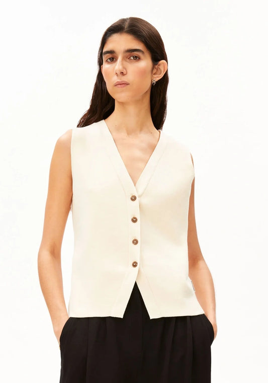 Woman wearing the ARMEDANGELS Canada Compact Buttoned Knit Vest in oatmilk with black pleated trousers, front view against a white background.