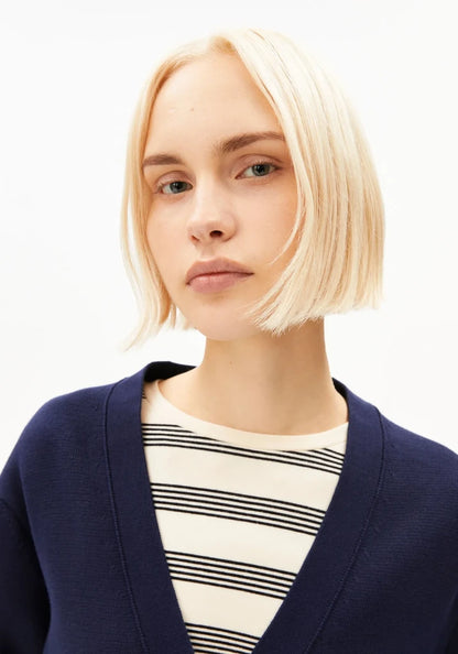 A woman with a short blonde bob wearing a navy blue cardigan over a cream and black striped shirt, photographed against a plain white background.