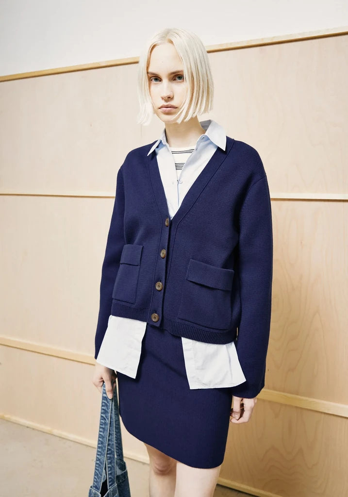 A blonde model wearing a navy blue cardigan and matching skirt over a white collared shirt, standing against a light wood-paneled wall.