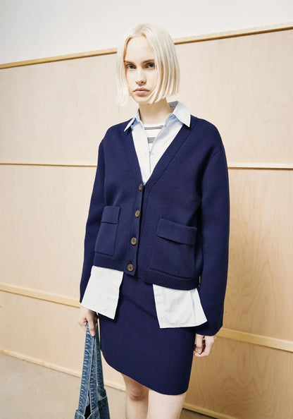 A blonde model wearing a navy blue cardigan and matching skirt over a white collared shirt, standing against a light wood-paneled wall.