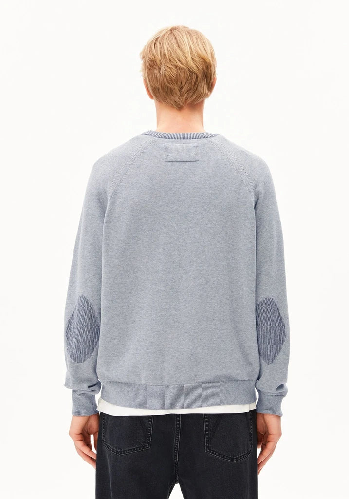 A man viewed from behind wearing a light gray crewneck sweatshirt with darker gray elbow patches, paired with dark jeans, against a plain white background.