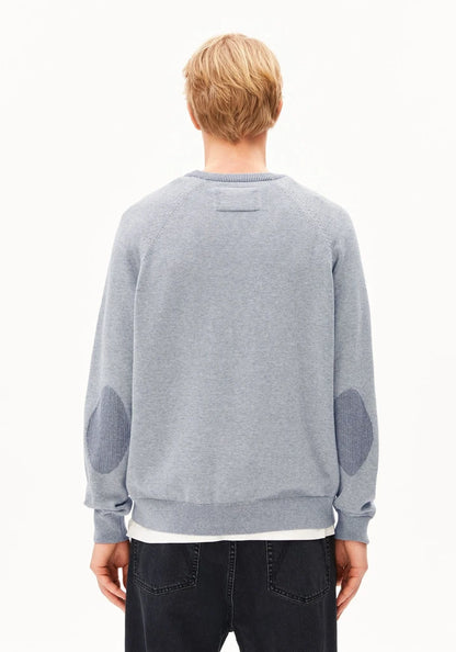 A man viewed from behind wearing a light gray crewneck sweatshirt with darker gray elbow patches, paired with dark jeans, against a plain white background.