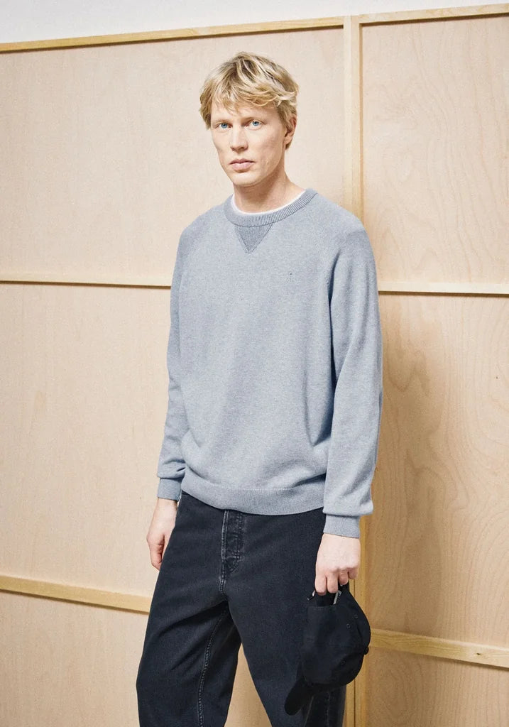 A man wearing a light gray crewneck sweater and dark jeans, holding a black cap, standing against a light wood paneled wall.
