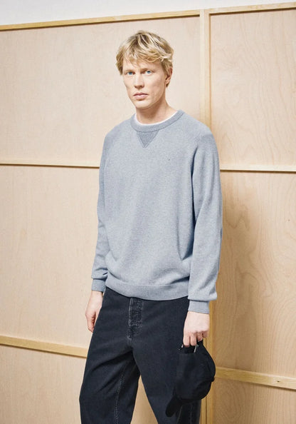 A man wearing a light gray crewneck sweater and dark jeans, holding a black cap, standing against a light wood paneled wall.