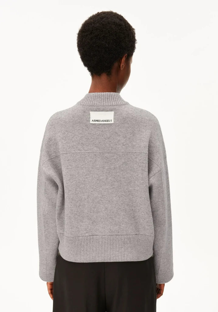 A person shown from the back wearing a light gray knit bomber jacket with a small white rectangular brand patch on the upper back, paired with dark pants, against a plain white studio background.