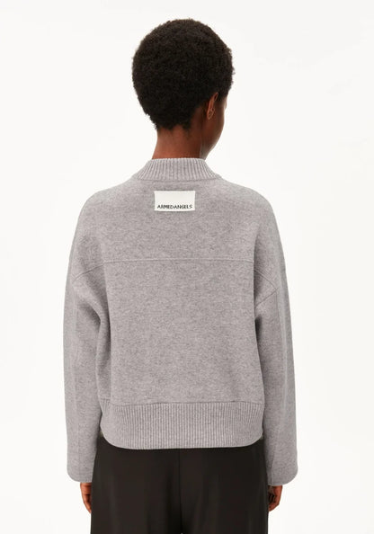 A person shown from the back wearing a light gray knit bomber jacket with a small white rectangular brand patch on the upper back, paired with dark pants, against a plain white studio background.