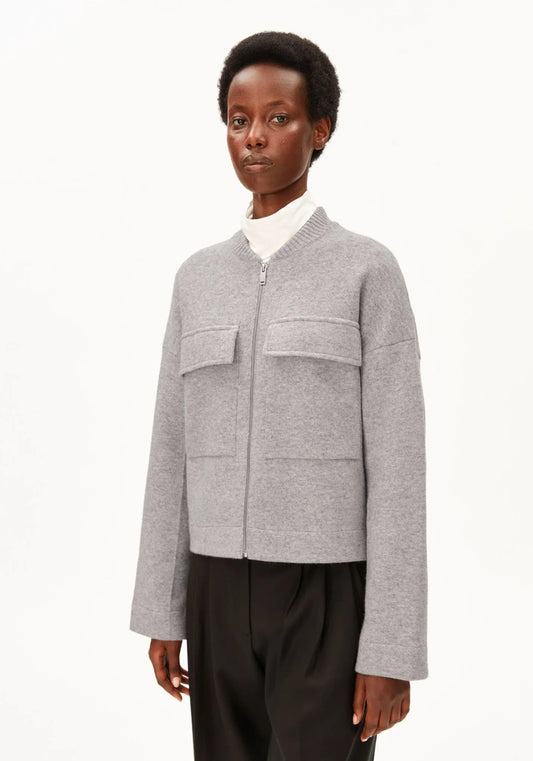 A model wearing a light gray zip-up knit bomber jacket with flap chest pockets, styled over a white turtleneck and black trousers, against a plain white studio background.