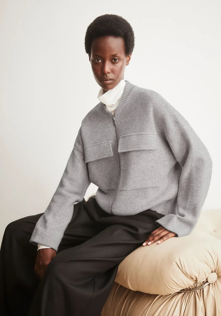 A model wearing a light gray zip-up knit bomber jacket with large front pockets, styled over a white mock neck top and dark wide-leg trousers, seated on a beige cushioned chair against a plain off-white background.