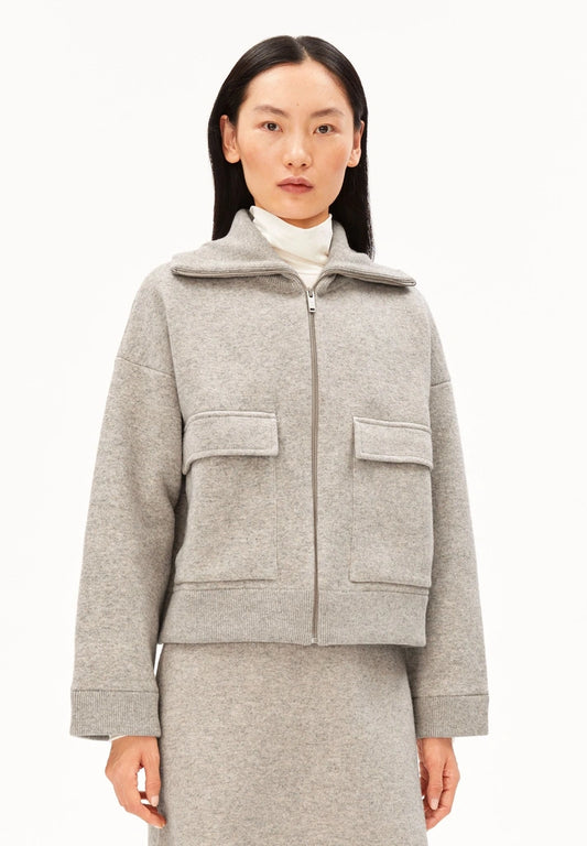 Woman wearing the ARMEDANGELS Canada Cotton-Wool Jacket in dark honey melange, zipped up over a white turtleneck, standing against a plain white studio background.