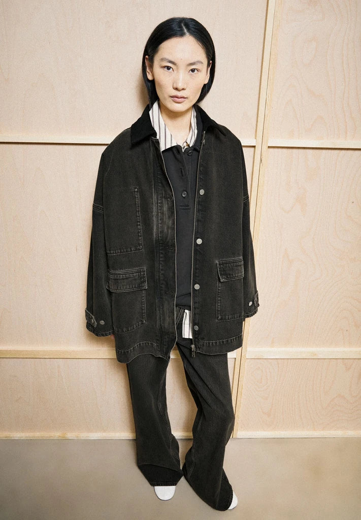 Woman wearing ARMEDANGELS Canada Denim Jacket Cord Collar with matching black jeans, standing in front of light wood panelling.