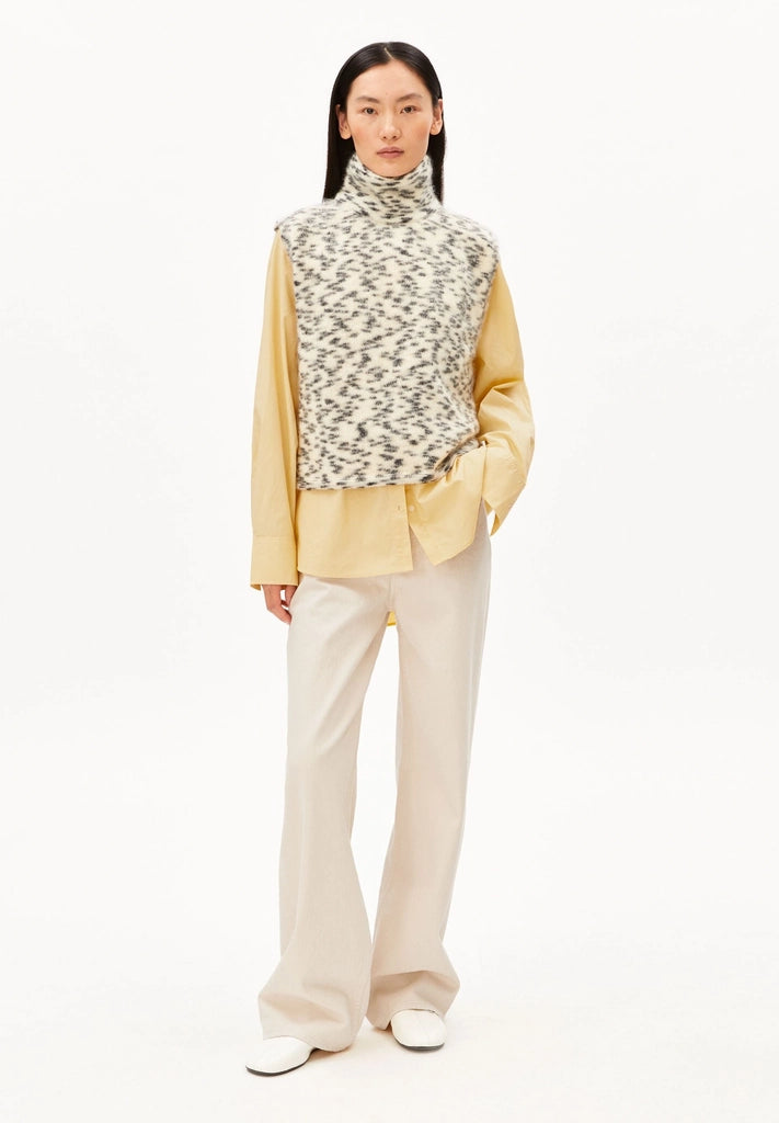 Full-length front view of a woman wearing ARMEDANGELS Canada Dotted Mohair-Blend Knit Vest in Oatmilk-Black with a yellow shirt, cream trousers and white sneakers, against a white background.