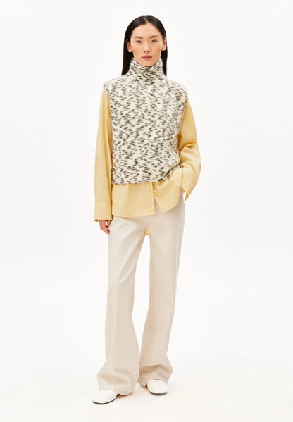 Full-length front view of a woman wearing ARMEDANGELS Canada Dotted Mohair-Blend Knit Vest in Oatmilk-Black with a yellow shirt, cream trousers and white sneakers, against a white background.