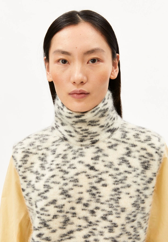 Close-up of a woman wearing ARMEDANGELS Canada Dotted Mohair-Blend Knit Vest in Oatmilk-Black, showing the mock neck and speckled knit, against a white background.