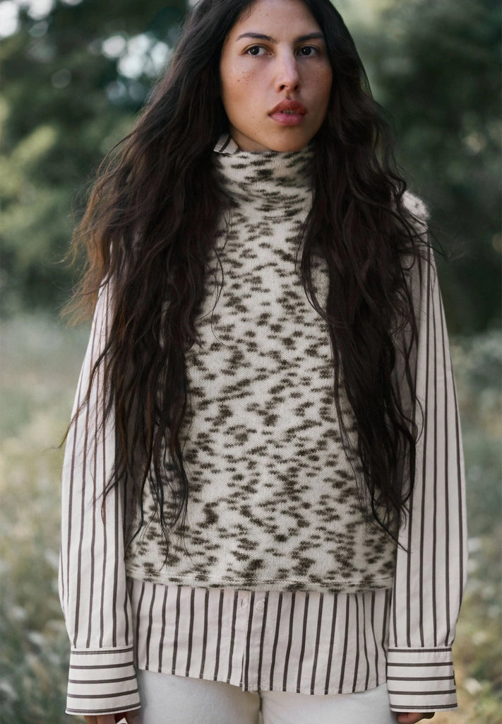 Woman wearing ARMEDANGELS Canada Dotted Mohair-Blend Knit Vest in Oatmilk-Black over a striped shirt, standing outdoors among greenery.