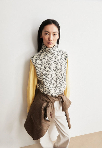 Woman wearing ARMEDANGELS Canada Dotted Mohair-Blend Knit Vest in Oatmilk-Black with a yellow shirt and a brown jacket tied at the waist, leaning against a light wall.