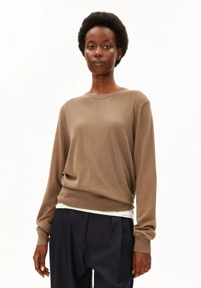 A model wearing a light brown crewneck knit sweater and dark navy trousers, photographed against a clean white studio background.