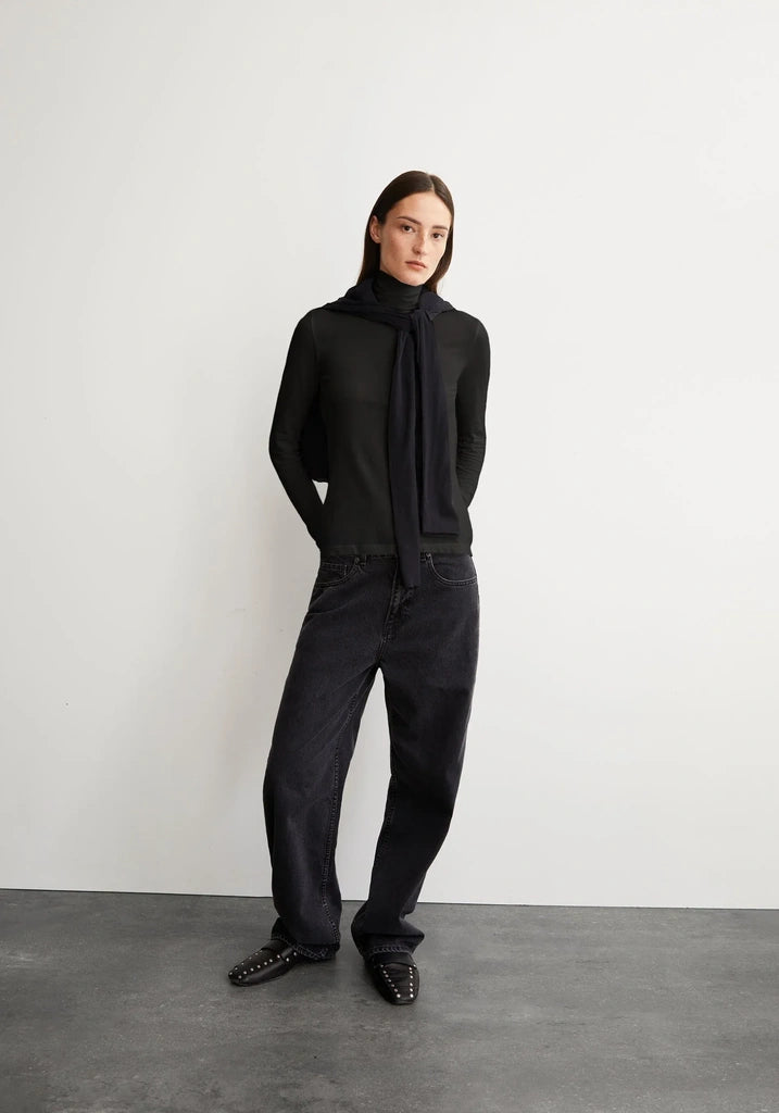 A model stands against a light gray wall and dark gray floor wearing a black knit mock-neck top with a long draped scarf, black wide-leg jeans, and black studded loafers.