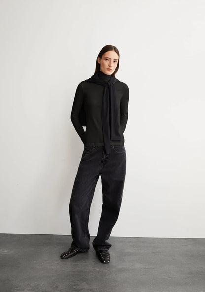 A model stands against a light gray wall and dark gray floor wearing a black knit mock-neck top with a long draped scarf, black wide-leg jeans, and black studded loafers.