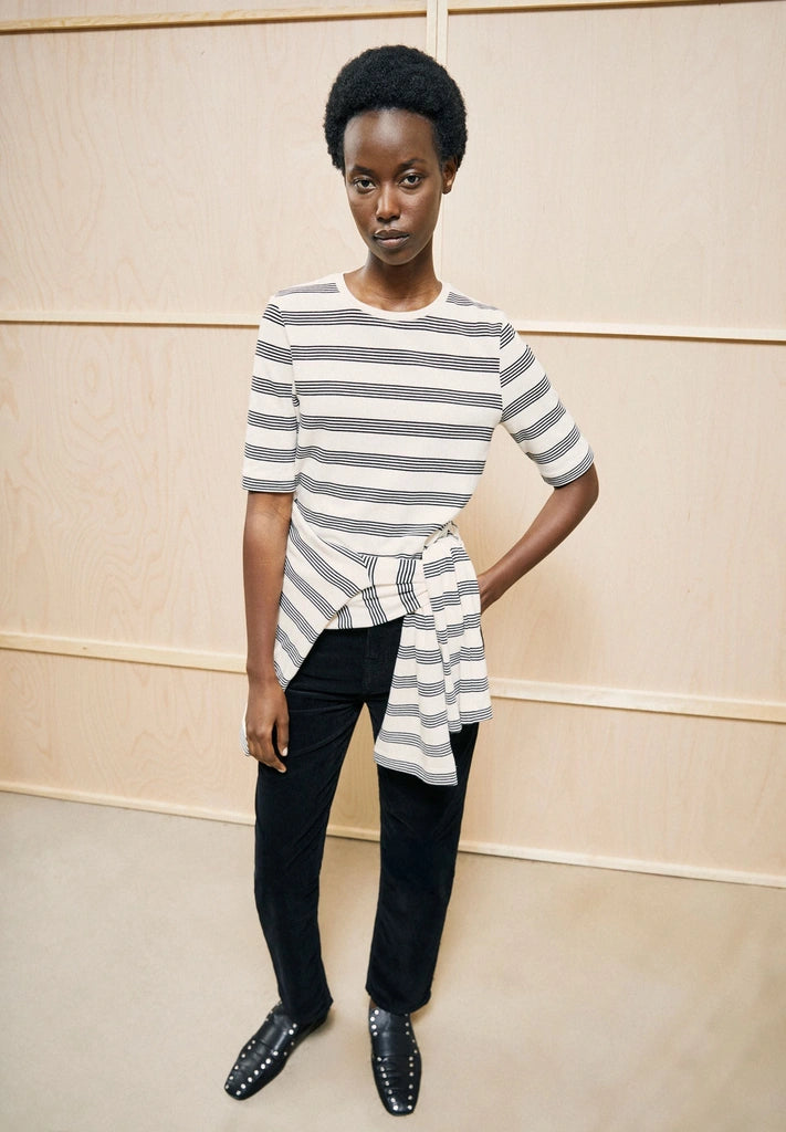 Woman wearing ARMEDANGELS Canada Jaanisara Fine Stripe T-Shirt in Oatmilk-Black with a striped shirt tied at the waist and black jeans, standing in front of light wood panelling.