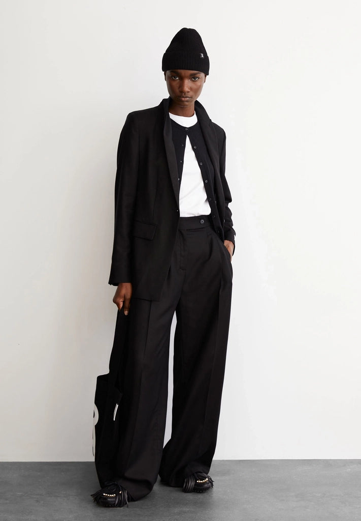 Woman wearing ARMEDANGELS Canada Kaayua Wide Leg Pants in black with a white tee, black blazer and black beanie, standing against a white wall on a grey floor.