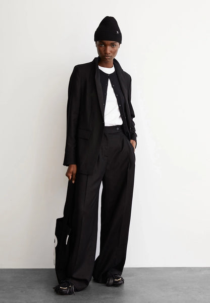 Woman wearing ARMEDANGELS Canada Kaayua Wide Leg Pants in black with a white tee, black blazer and black beanie, standing against a white wall on a grey floor.